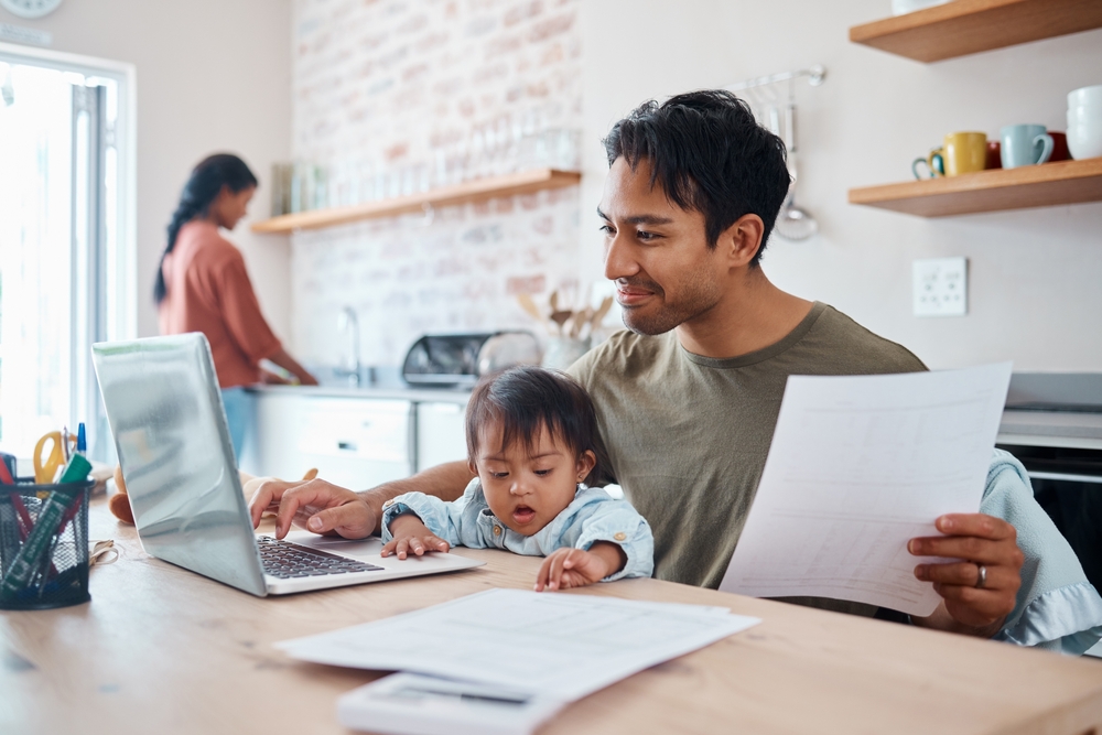 Man sitting in kitchen looking at laptop with baby on his lap.